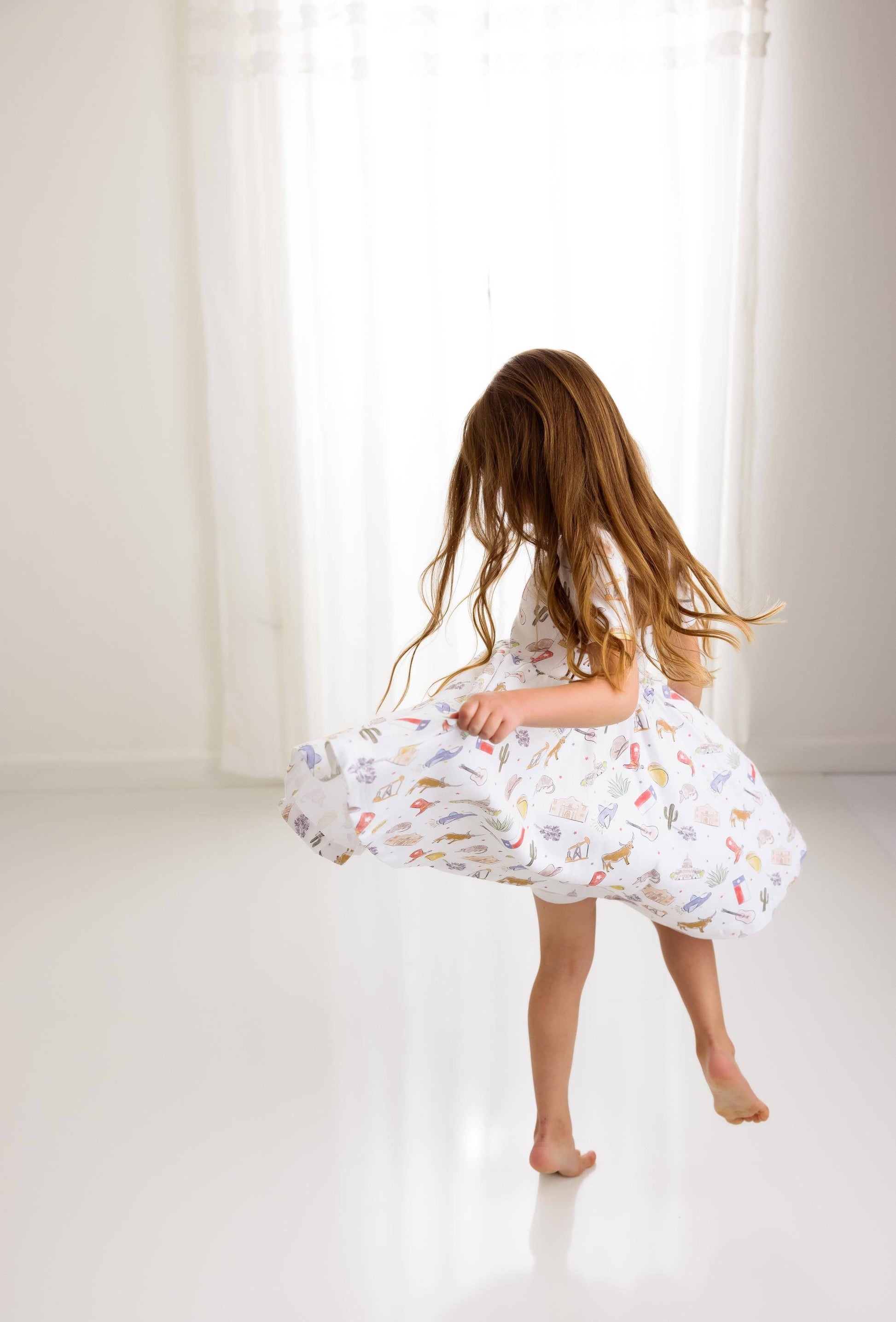 Young girl in a white dress with colorful Texas patterns standing in a bright room.