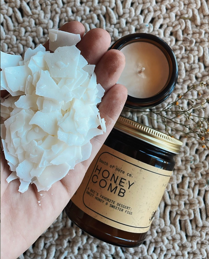 Hand holding a jar of 'Honey Comb' candle with a lit candle and honeycomb pieces on a textured surface.