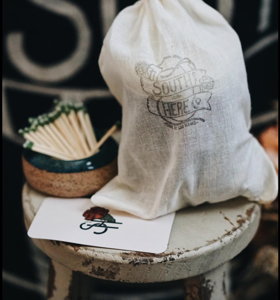 White drawstring bag with 'Southern in Here' branding on a rustic surface with matches and a card.