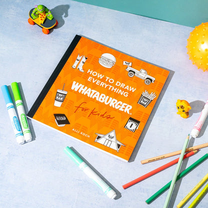 Book titled 'How to Draw Everything Whataburger for Kids' on a light blue surface with drawing tools.