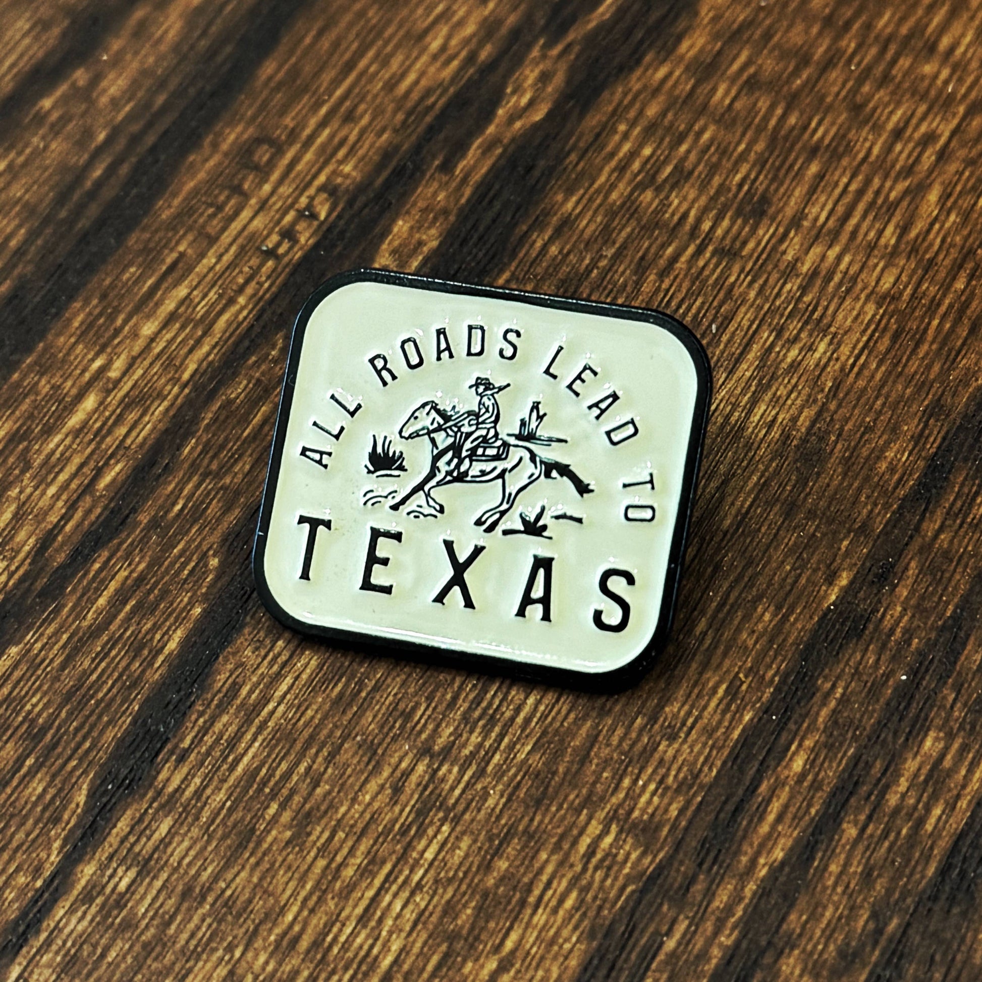 Enamel pin with 'All Roads Lead to Texas' design on a wooden surface