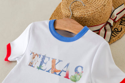 White t-shirt with colorful text and graphics on a wooden hanger, with a straw hat and scarf in the background.